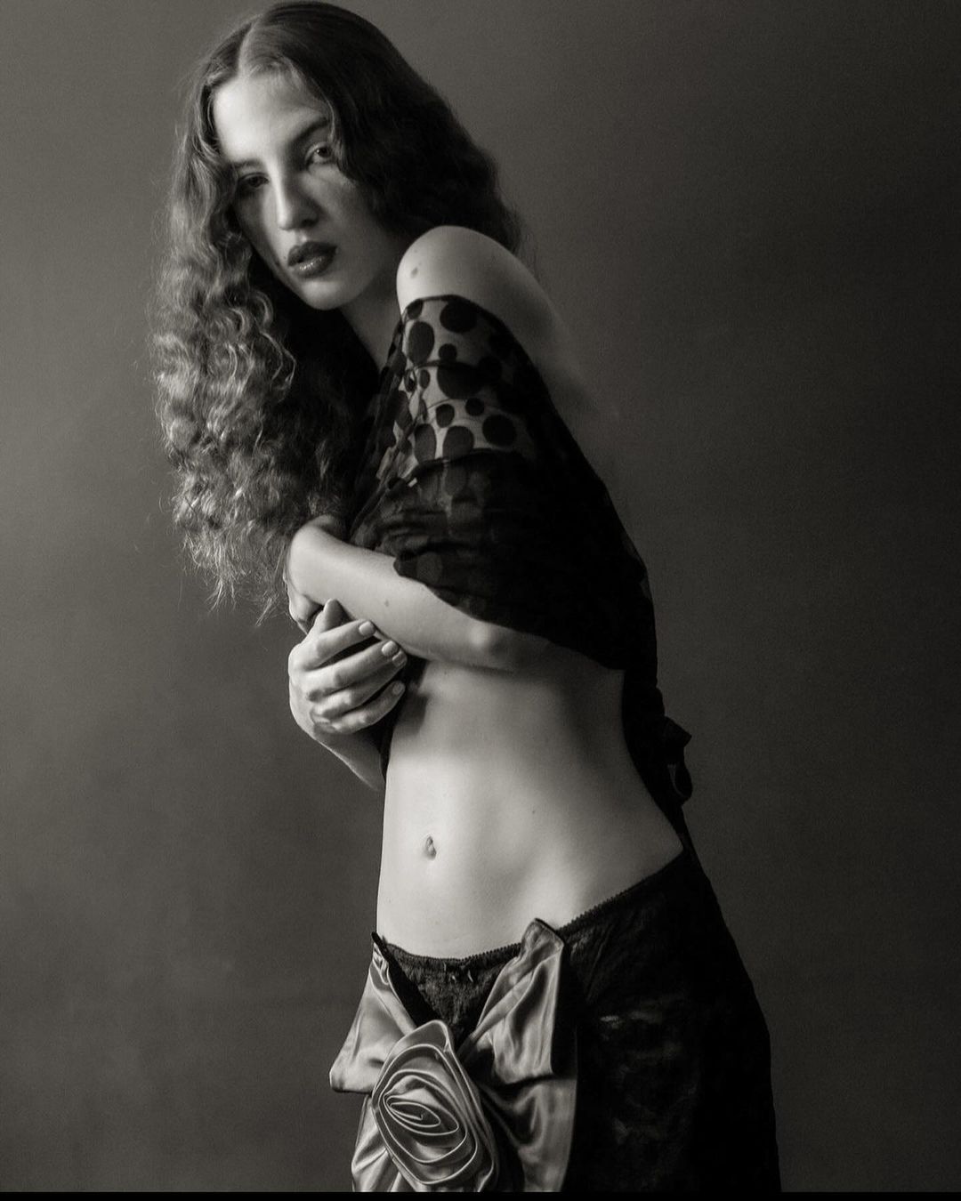 Monochrome portrait of a woman with long curly hair, wearing black lace lingerie and a polka dot shawl, against a dark background.