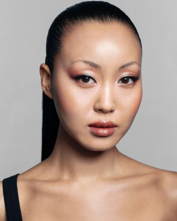 Close-up beauty portrait of an East Asian woman with sleek dark hair pulled back against a grey backdrop.
