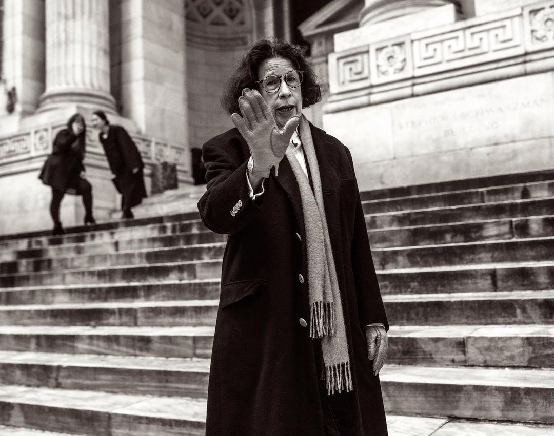 A woman in a coat and glasses stands on stone steps, raising a gloved hand, with two blurry figures visible in the background.