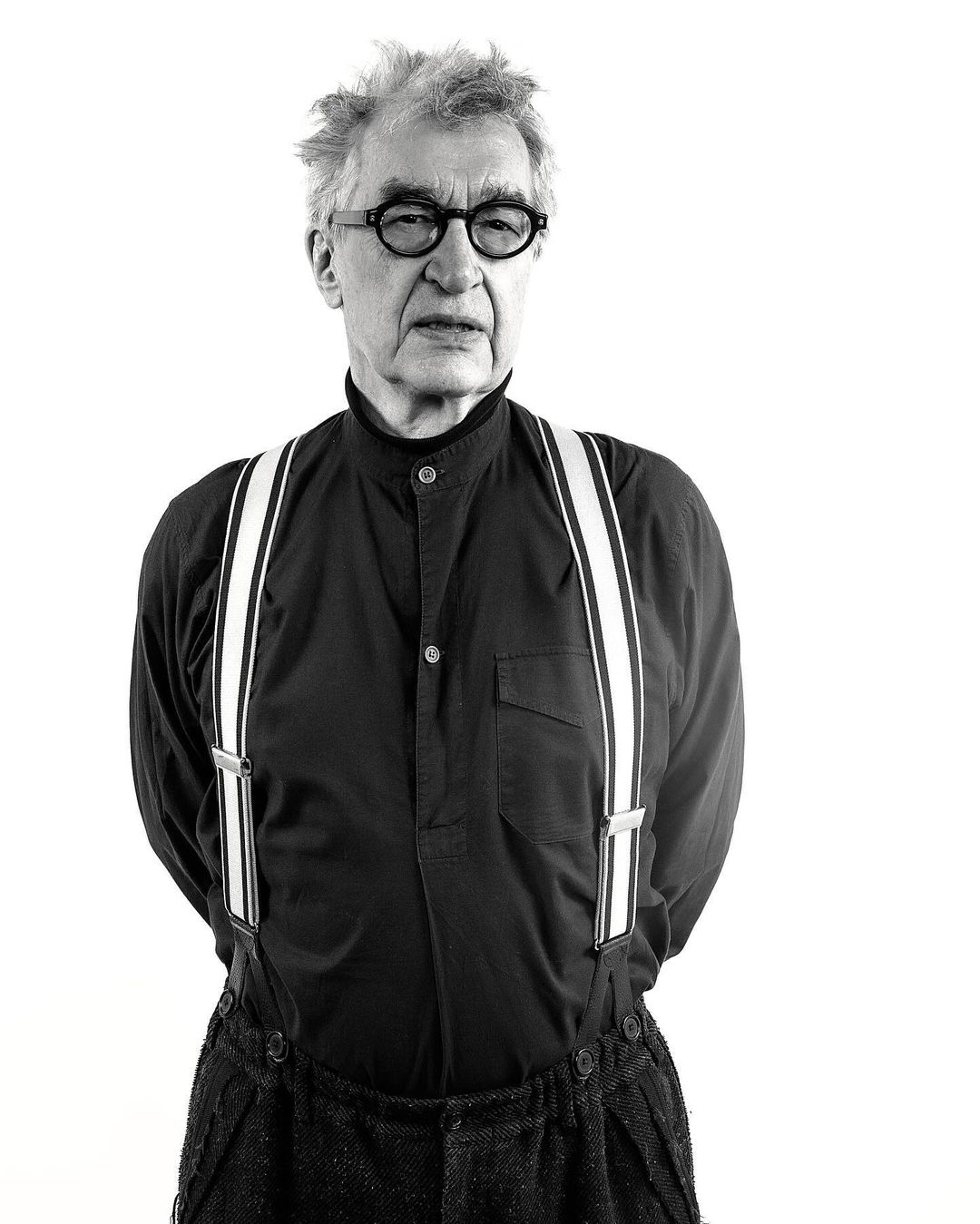Black and white portrait of an older adult male wearing a dark shirt, suspenders, and glasses, set against a bright white background.