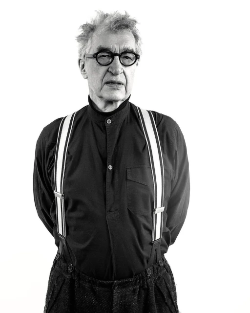 Black and white portrait of an older adult male wearing a dark shirt, suspenders, and glasses, set against a bright white background.