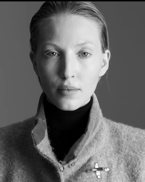 Monochrome close-up portrait of a young woman with light skin, wearing a dark turtleneck and a light-colored coat with a metal clasp.