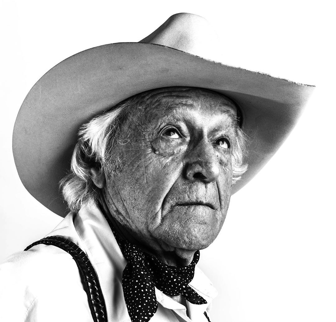 Black and white portrait of an elderly man wearing a cowboy hat, suspenders, and a patterned scarf against a white background.