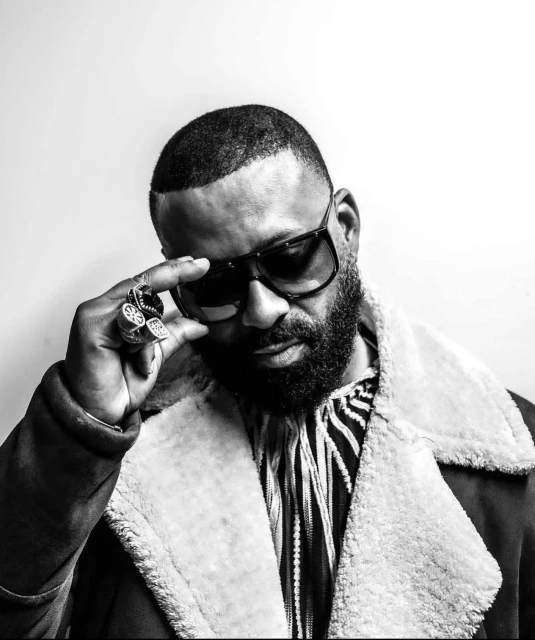 Black and white portrait of an adult male with a beard and sunglasses, wearing a patterned shirt and coat, against a white background.