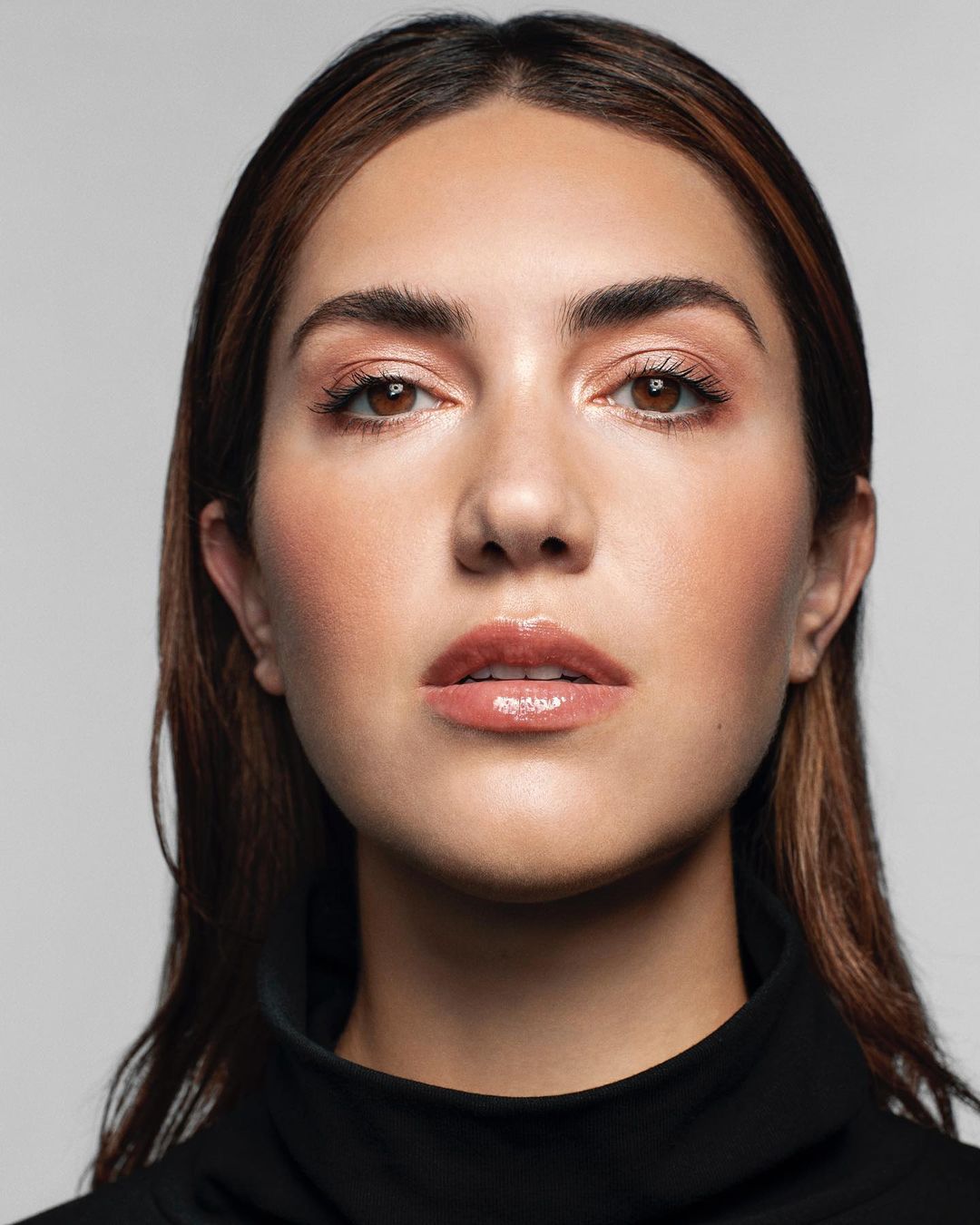 Close-up portrait of a brunette woman with glossy lip makeup, wearing a black top, set against a neutral grey backdrop in a studio setting.