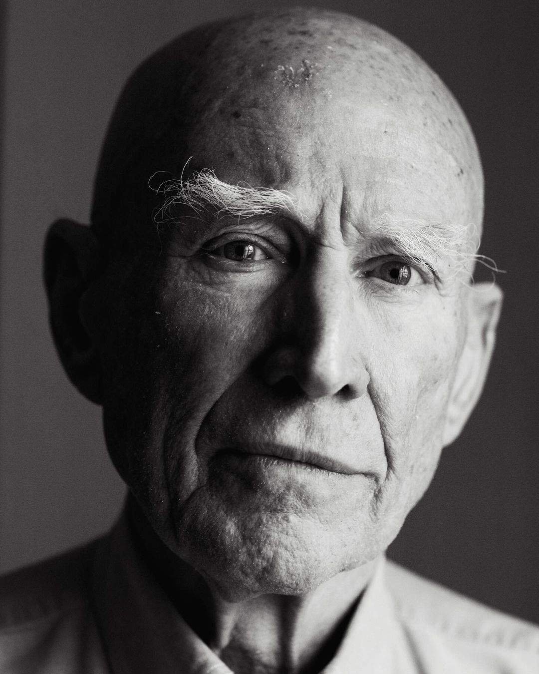 Black and white portrait of an elderly man with a bald head, light eyes, and furrowed brow, captured in a studio setting with soft lighting.