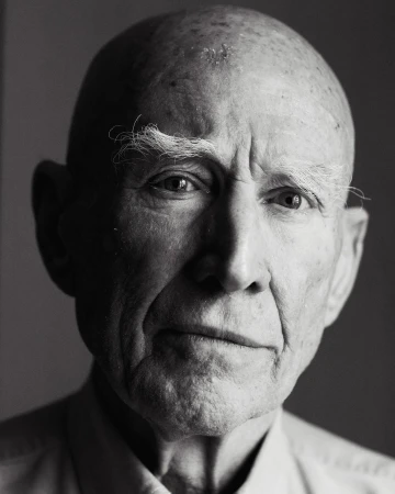 Black and white portrait of an elderly man with a bald head, light eyes, and furrowed brow, captured in a studio setting with soft lighting.