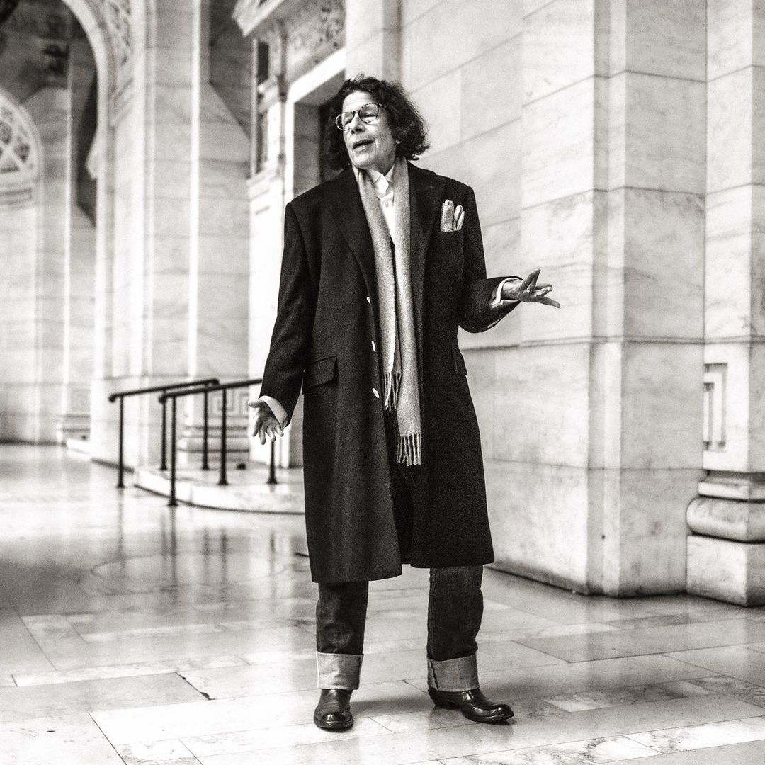 Black and white full shot of an adult male standing and gesturing in a marble building hall wearing a coat, scarf, and dark trousers.