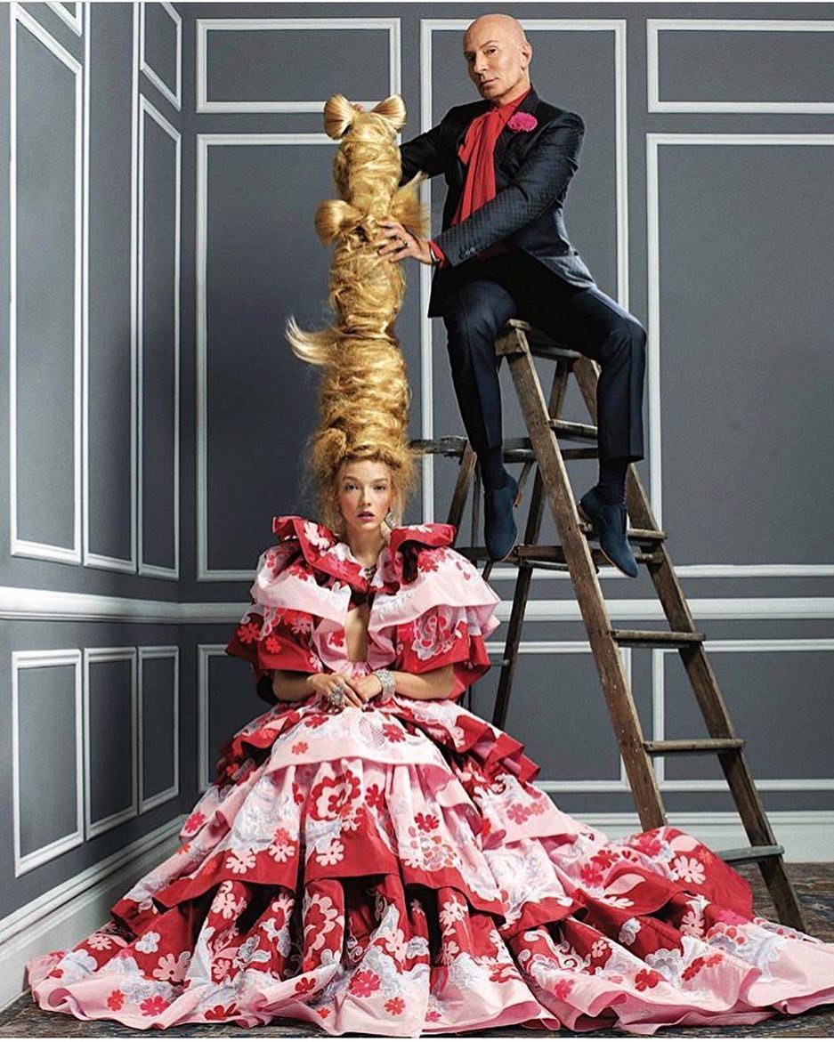 Fashion shot featuring a model in a red and pink floral gown with a large, sculpted hairstyle, and a man on a ladder, indoor studio.