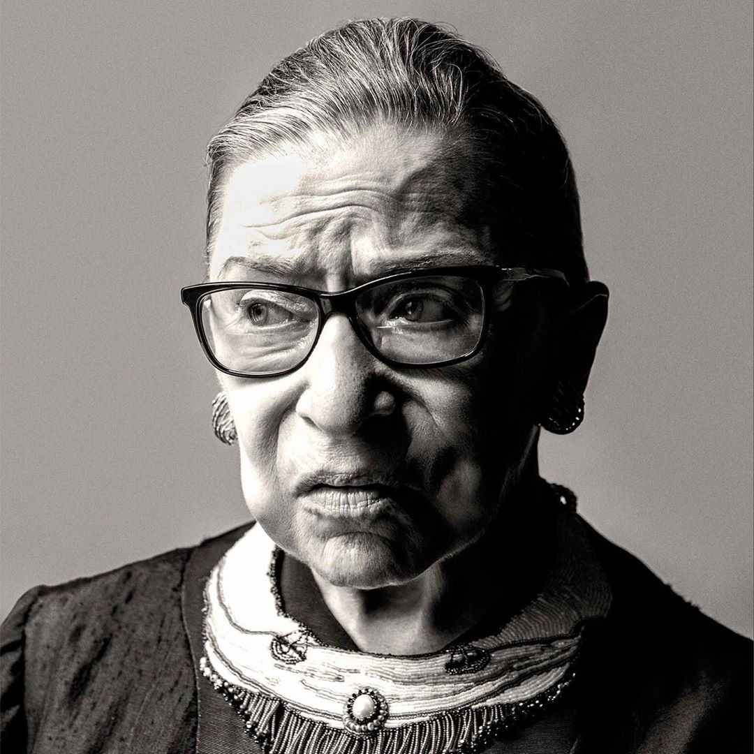 Black-and-white portrait of Ruth Bader Ginsburg with eyeglasses and necklace, gazing slightly to the left in a studio setting.