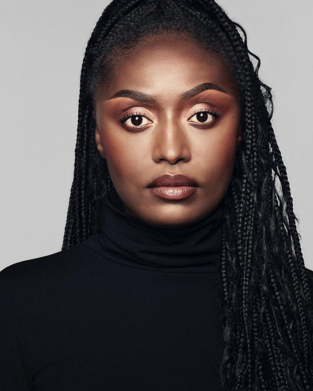 Portrait of a confident Black woman with braided hair, wearing a black turtleneck sweater against a neutral gray background.