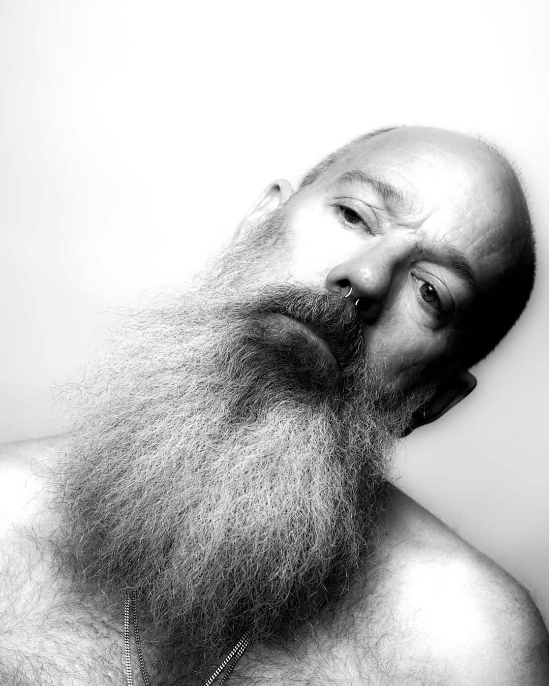 Black and white portrait of a bald adult male with a full, long beard, nose ring, and chains. He is looking at the camera. 