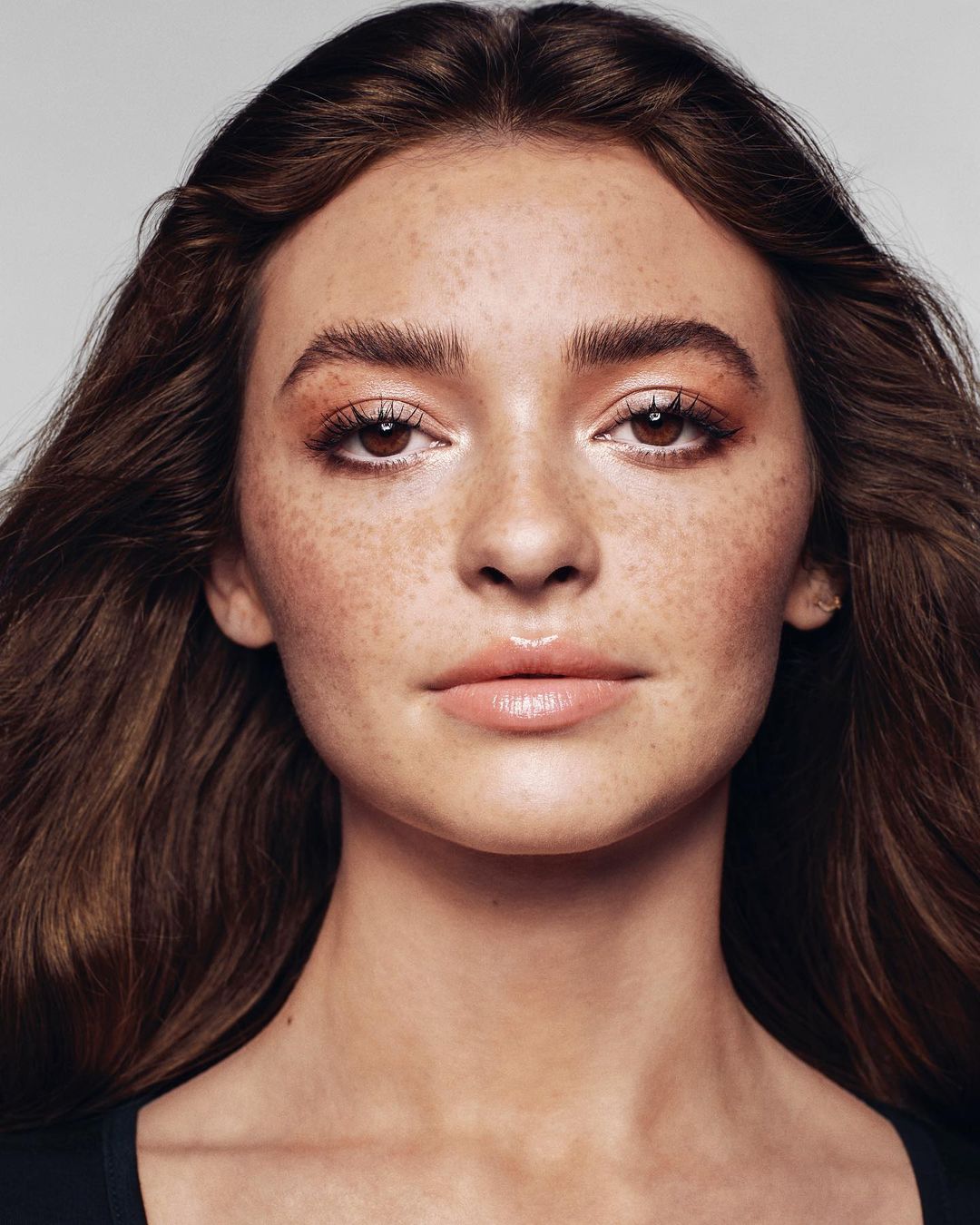 Close-up portrait of a young adult female with freckles and wavy brown hair against a neutral grey background in a studio setting.