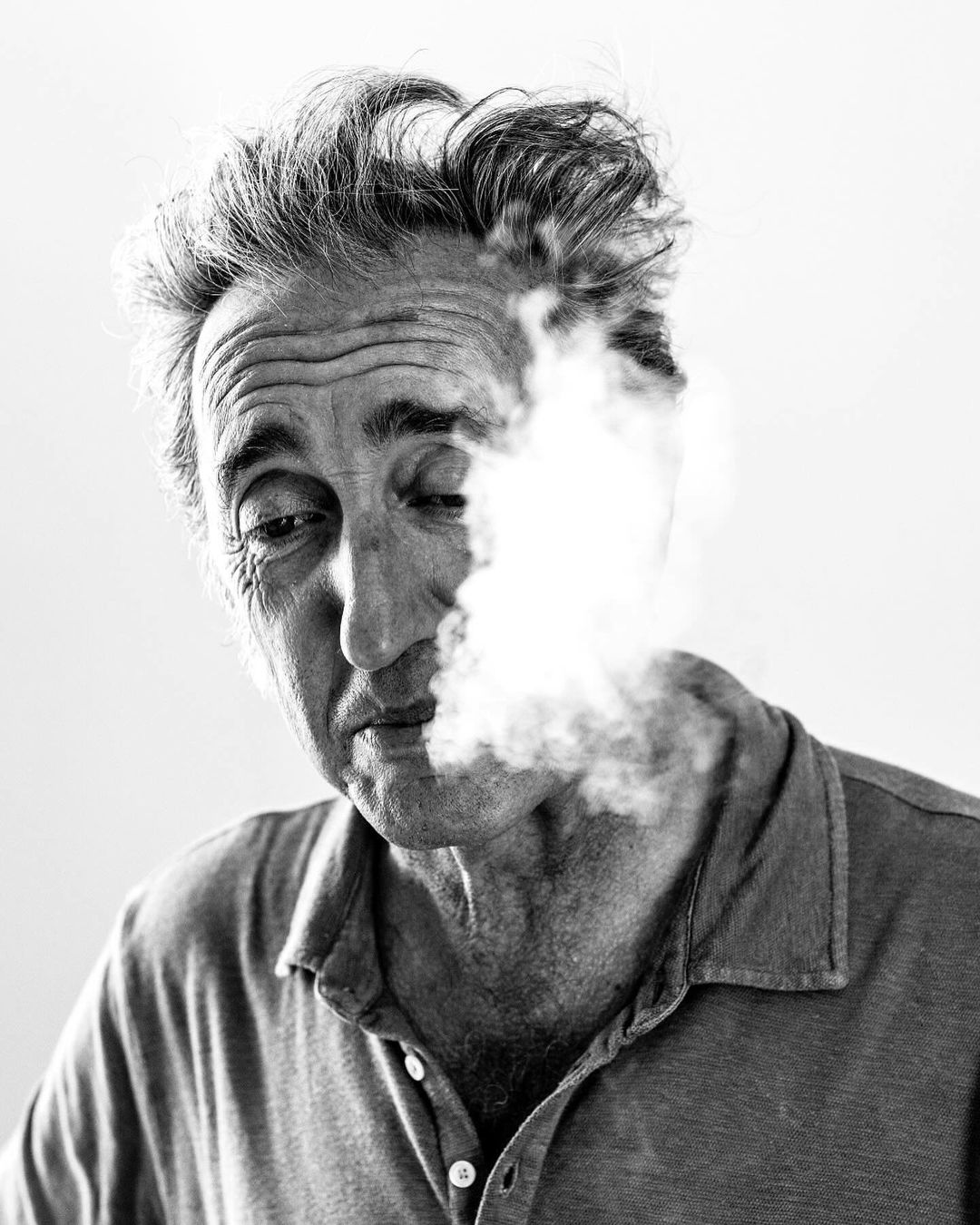 Black and white portrait of a middle-aged man with messy hair, smoking. White smoke is billowing around his face and neck.