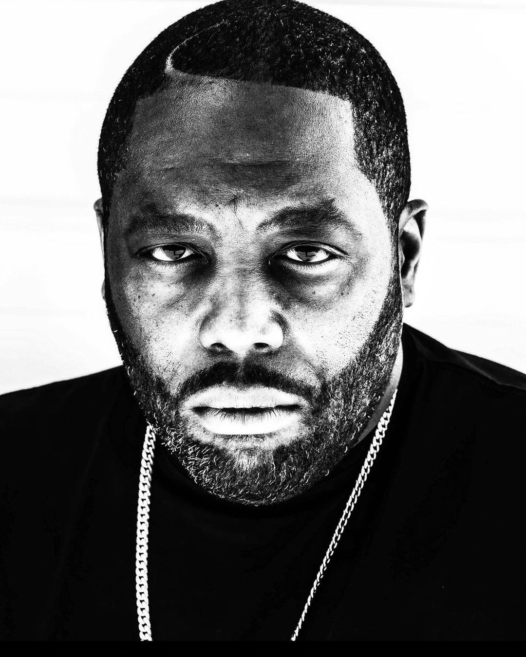 Monochrome portrait of an adult male with a beard, wearing a chain necklace, against a bright background.