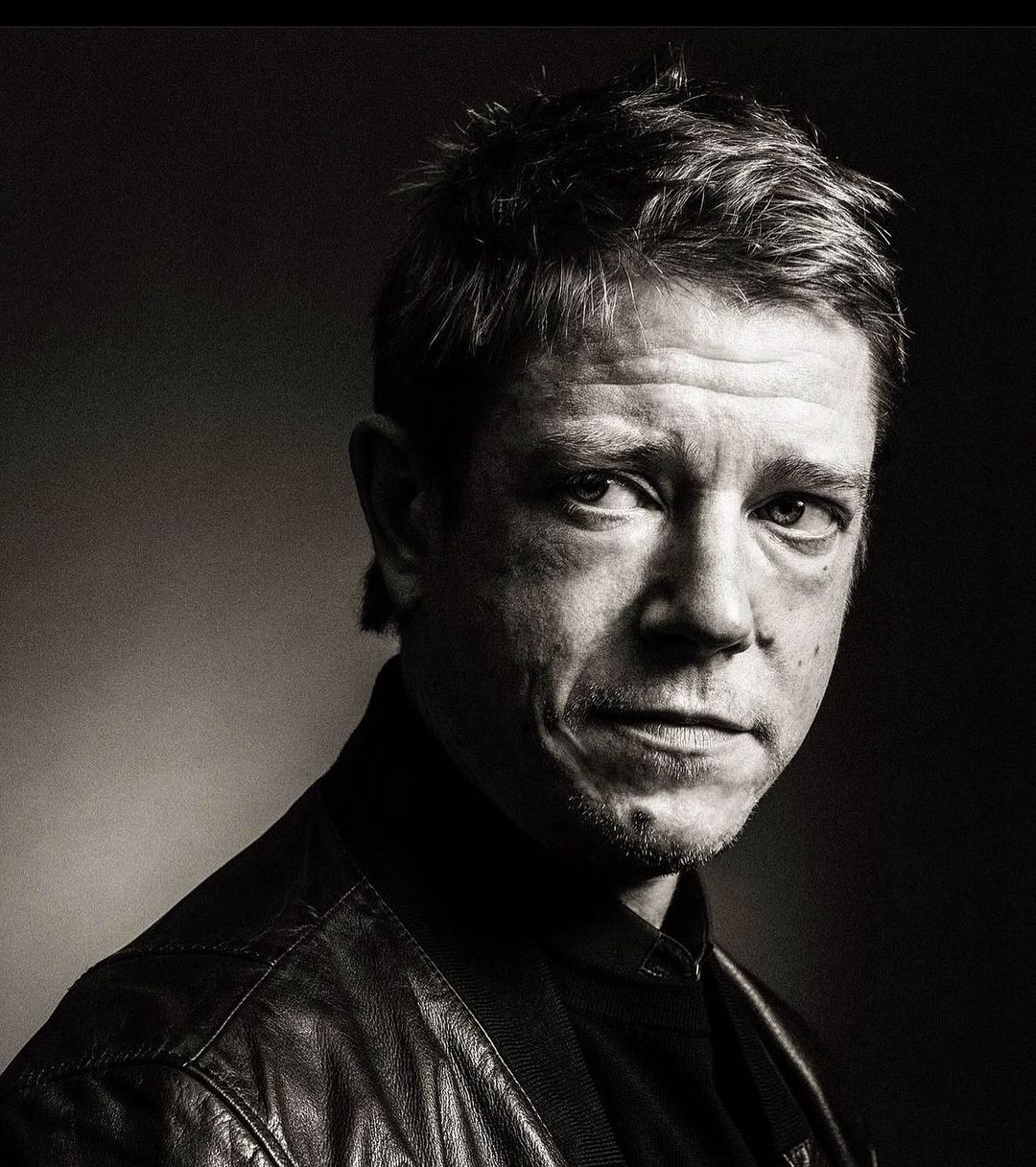 Monochrome portrait of a man in his 40s wearing a dark leather jacket against a dark background