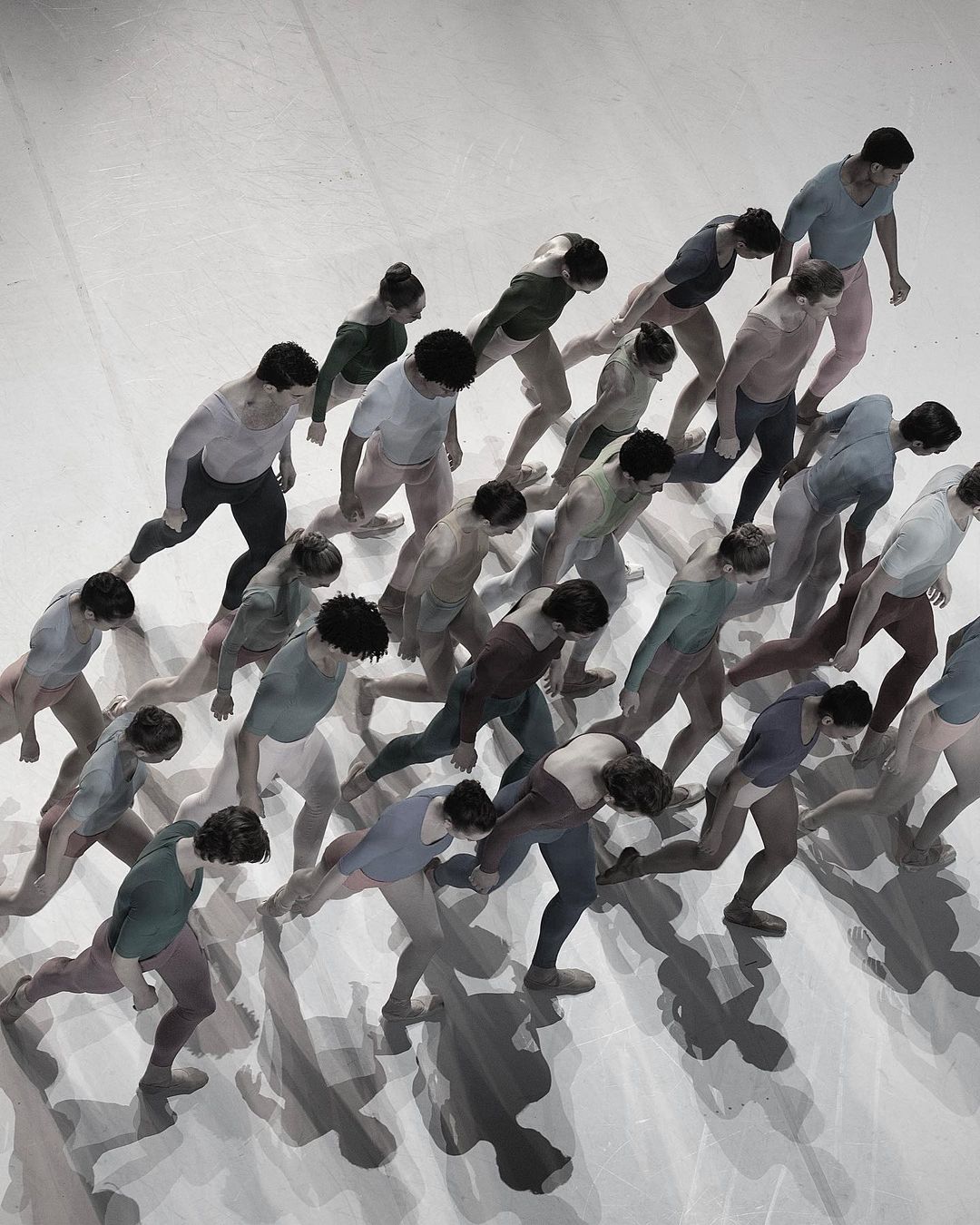 Overhead shot of a group of adult dancers in colorful clothing, moving in unison across a white floor, their shadows trailing behind them.