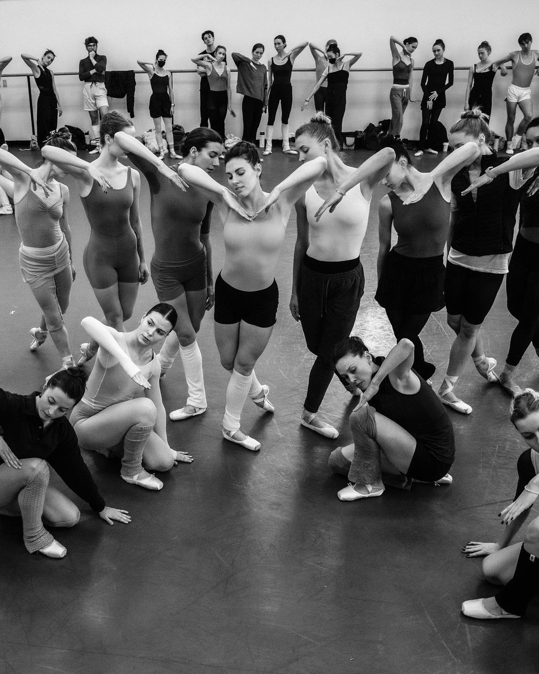 Group of ballet dancers in leotards and tights practice in a studio, some seated, some standing, with arms extended and legs posed.