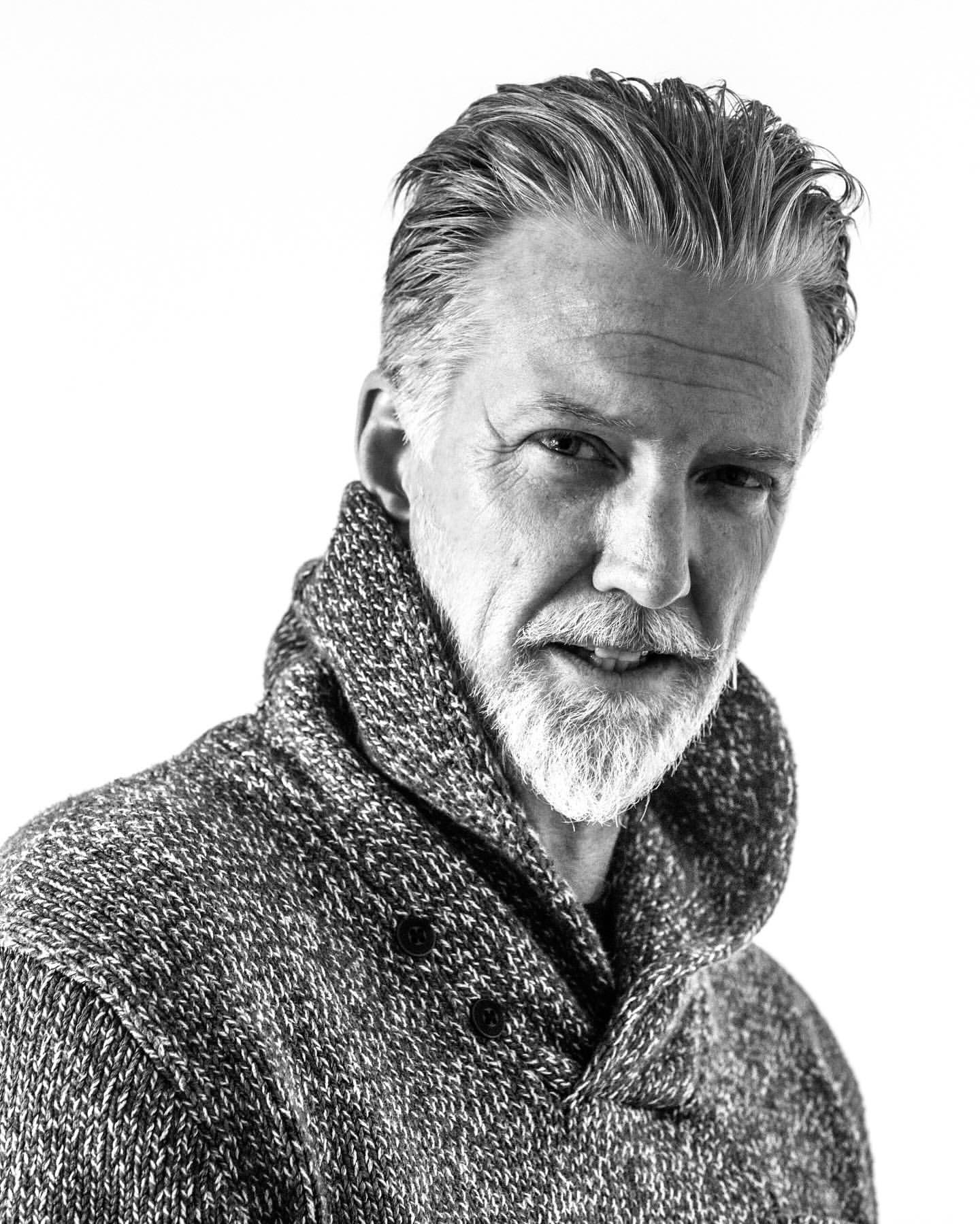 Black and white portrait of a mature adult man with a beard and gray hair wearing a knitted sweater against a bright, neutral background.