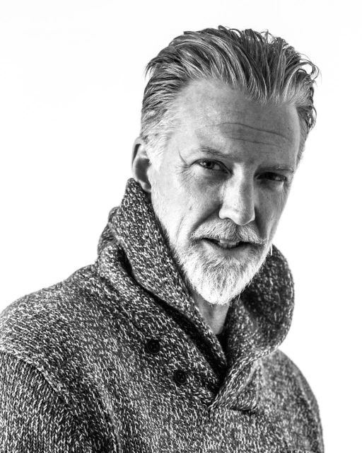 Black and white portrait of a mature adult man with a beard and gray hair wearing a knitted sweater against a bright, neutral background.
