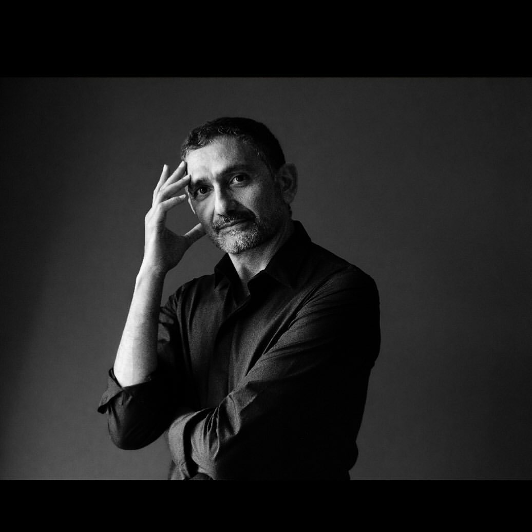 Black and white portrait of a man with short dark hair and a beard, wearing a dark collared shirt, posing with his hand on his head.