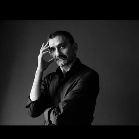 Black and white portrait of a man with short dark hair and a beard, wearing a dark collared shirt, posing with his hand on his head.