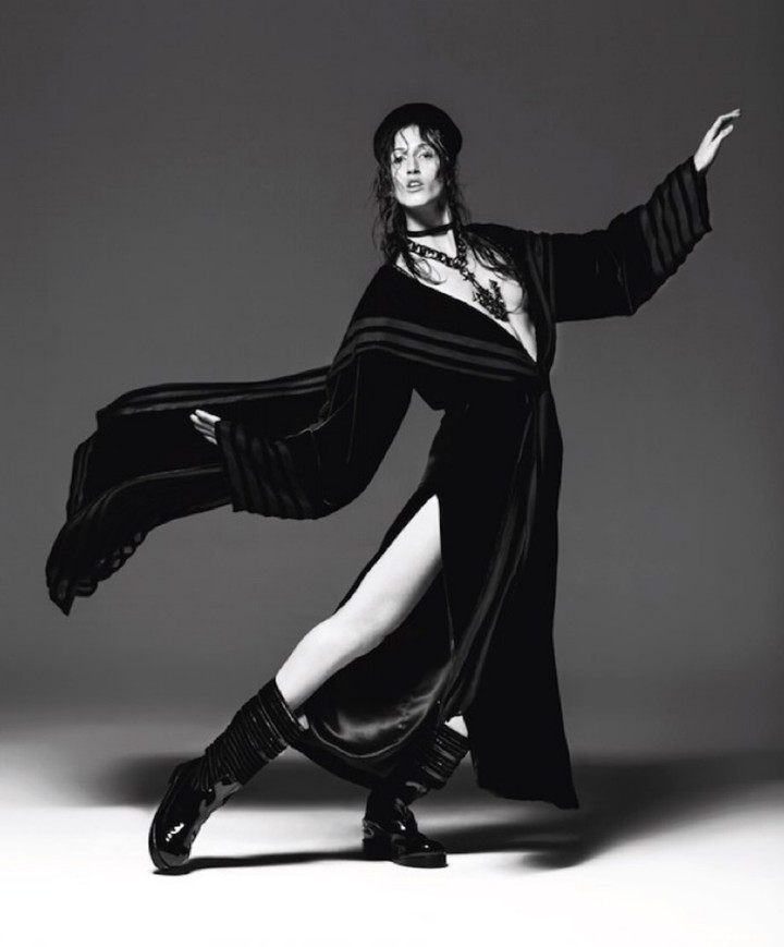A stylish woman poses in a black dress, shiny black boots, and hat against a grey background, showcasing a blend of elegance and edge.