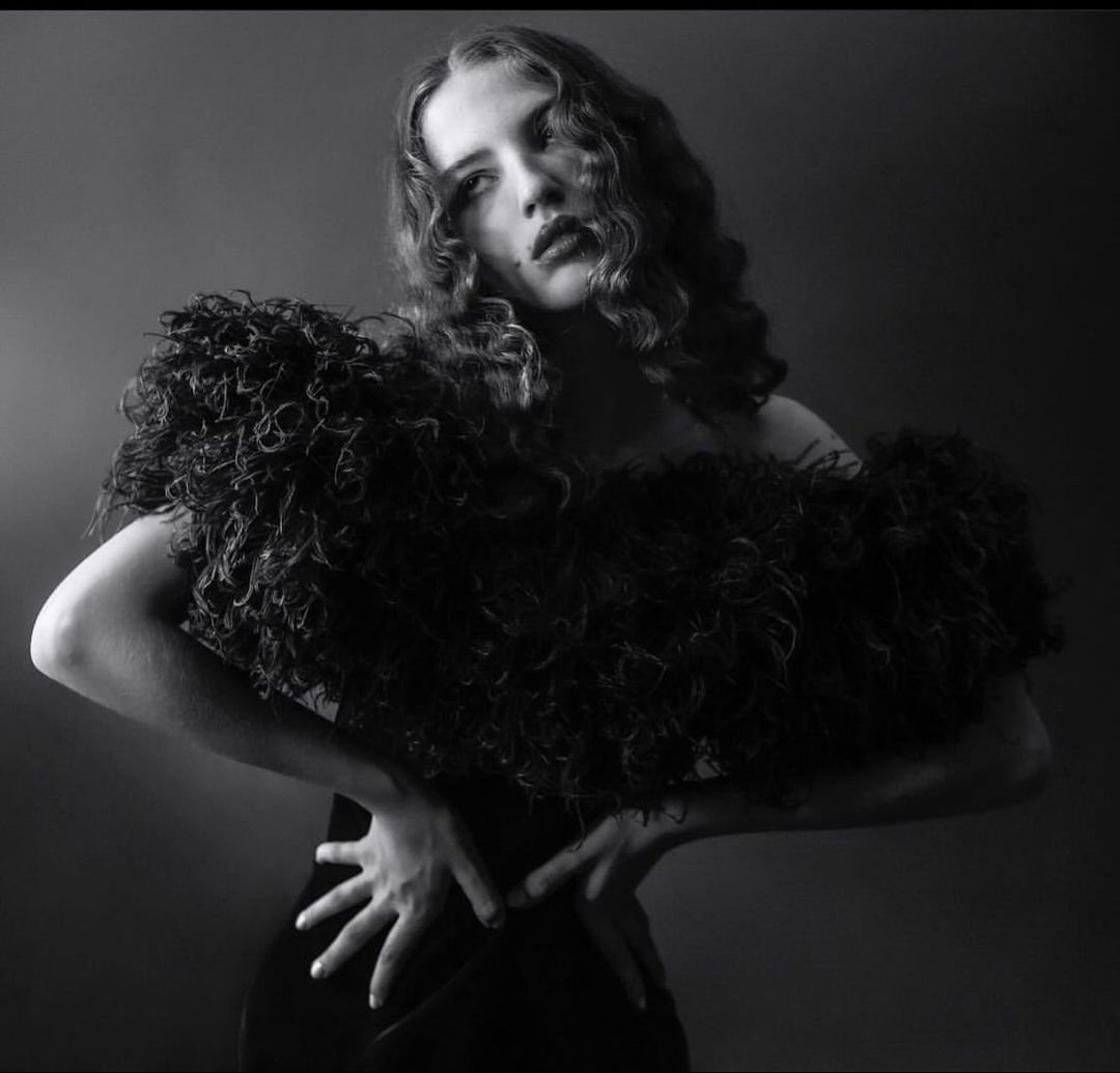 Black and white portrait of a woman with curly hair wearing an ornate ruffled top, set against a dark, neutral background.