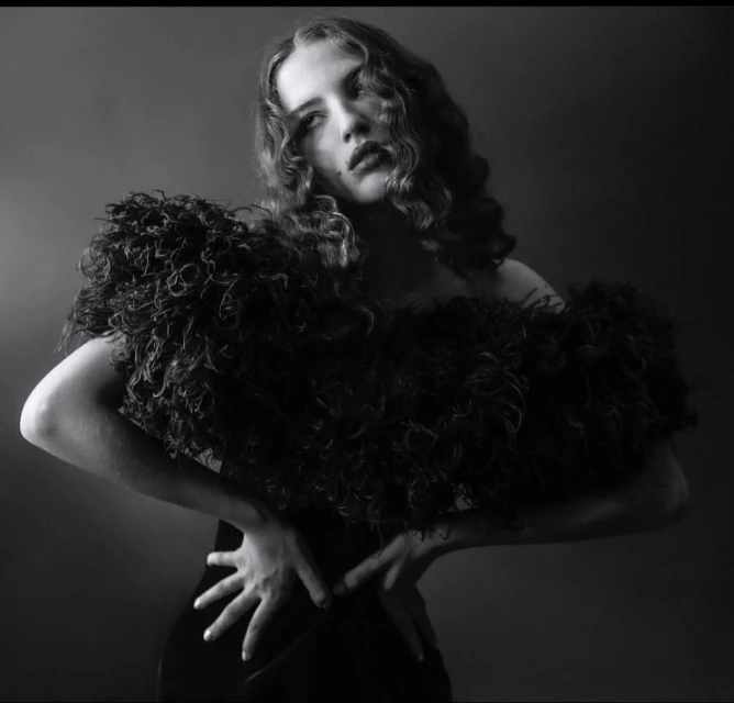 Black and white portrait of a woman with curly hair wearing an ornate ruffled top, set against a dark, neutral background.