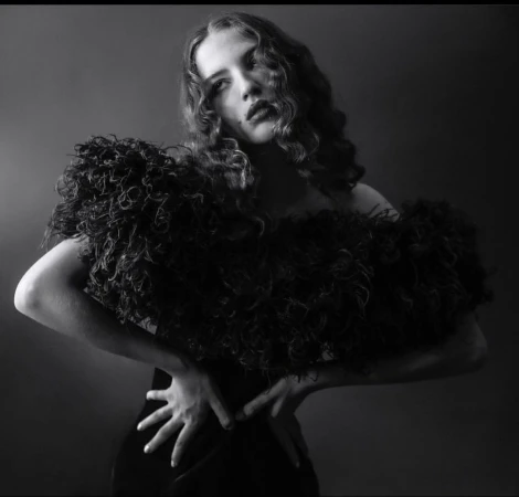 Black and white portrait of a woman with curly hair wearing an ornate ruffled top, set against a dark, neutral background.