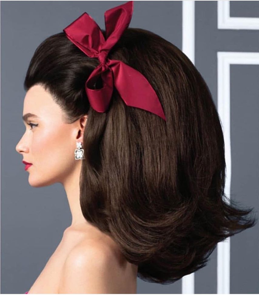 Profile of a woman with dark brown voluminous hair, styled with a red bow, and wearing elegant, dangling earrings against a gray backdrop.