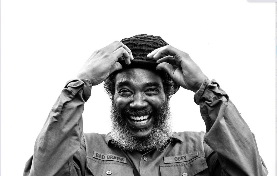 Black and white portrait of a Black adult male smiling with a cap on his head, wearing a collared shirt, in front of a white background.