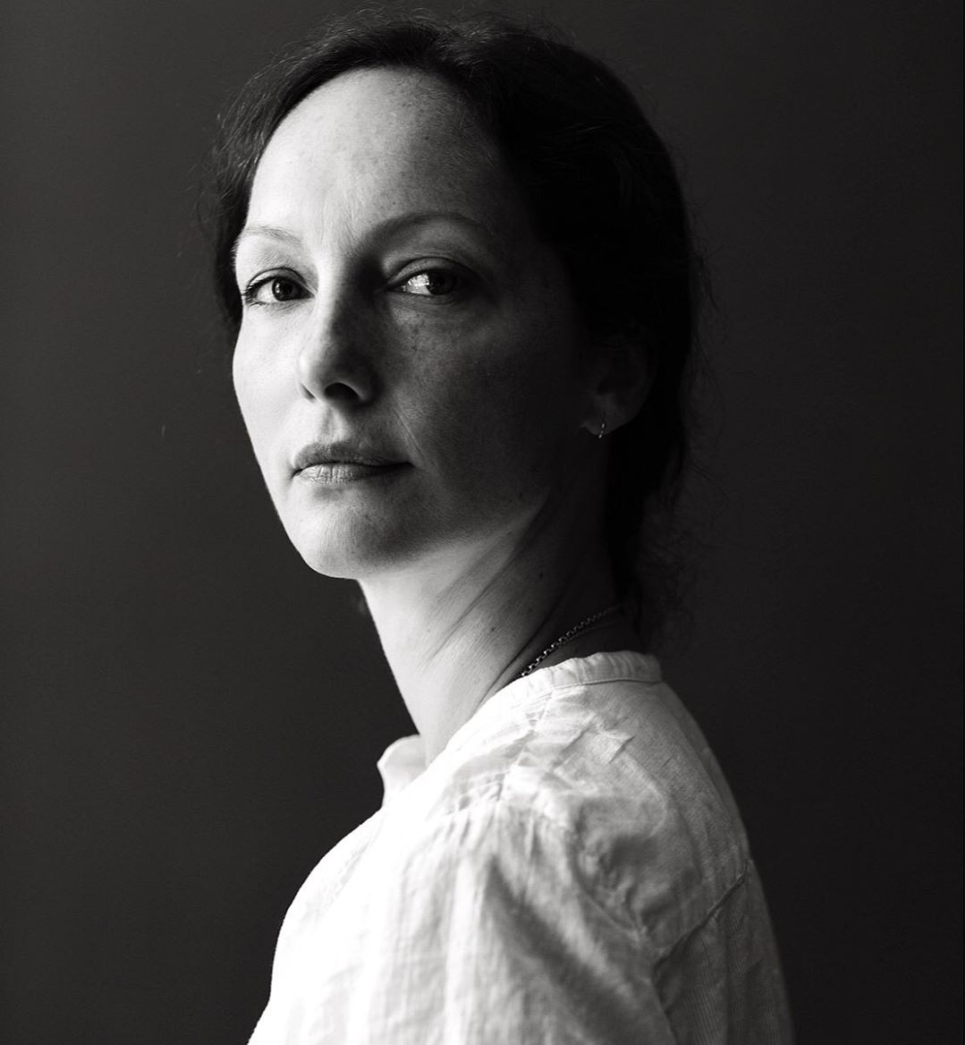 Monochrome portrait of an adult woman with dark hair looking at the camera, wearing a collared shirt against a dark background.