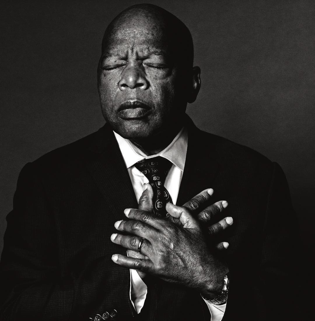 Black and white portrait of an adult male with hands clasped over his chest, eyes closed, wearing a suit and tie against a dark background.