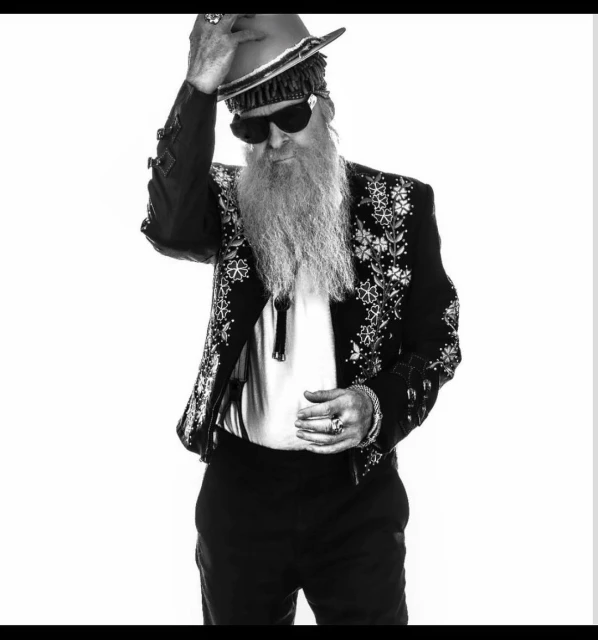 Monochrome portrait of bearded male musician in sunglasses and decorative jacket tipping western hat on white background in studio.