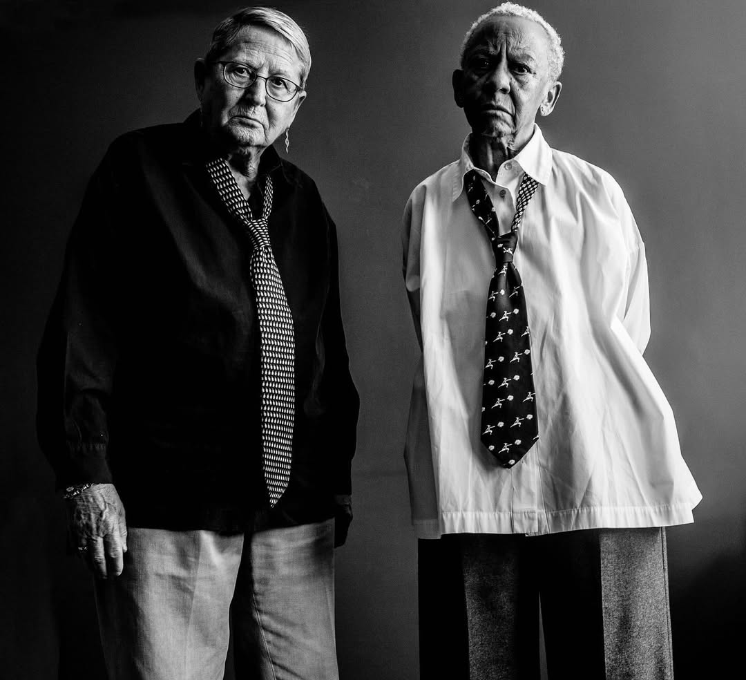Black and white portrait of two elderly women wearing ties and collared shirts in a photo studio setting, looking directly at the camera.