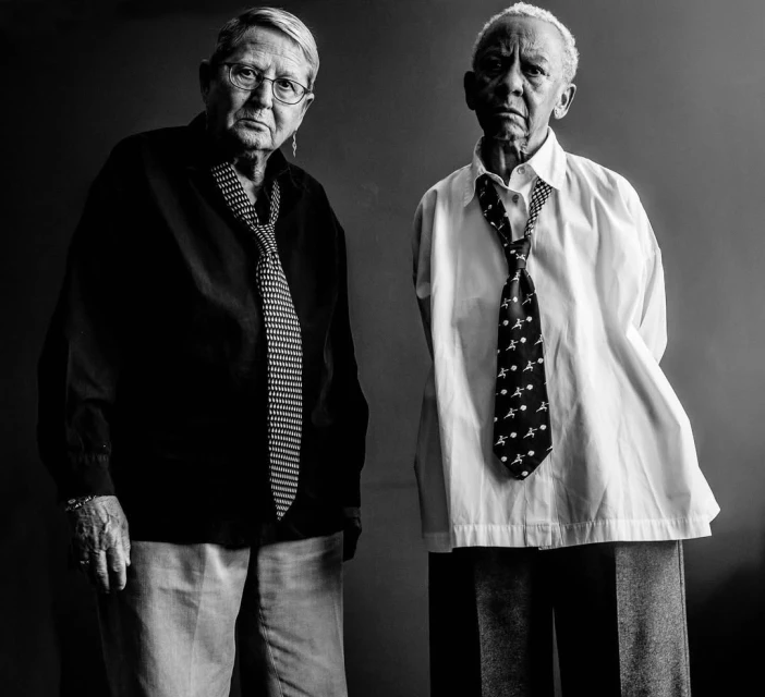 Black and white portrait of two elderly women wearing ties and collared shirts in a photo studio setting, looking directly at the camera.