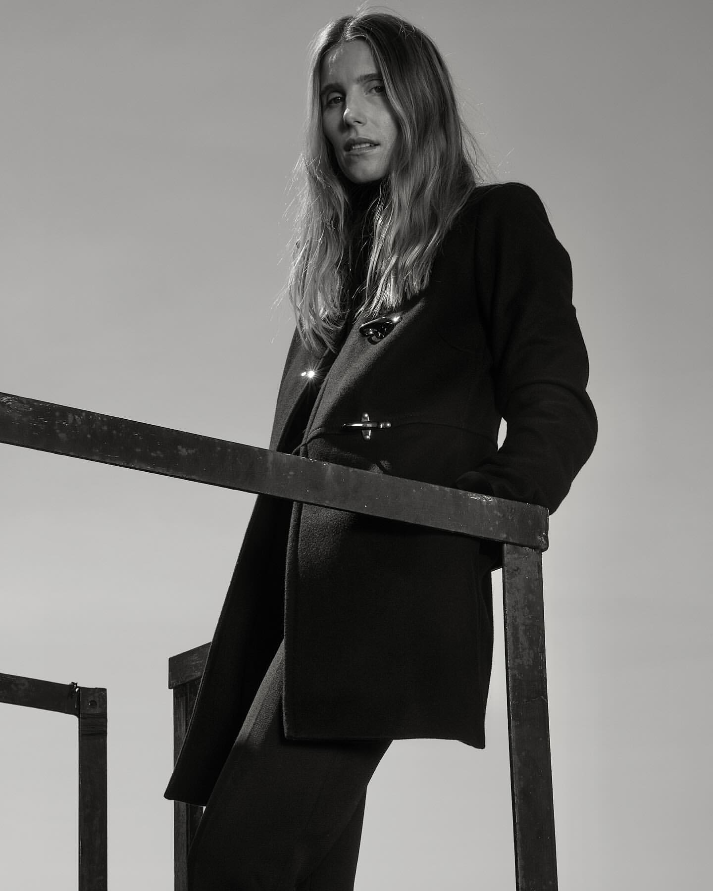 Monochrome portrait of an adult female in a dark coat standing on a black metal structure against a light backdrop in a fashion editorial style.