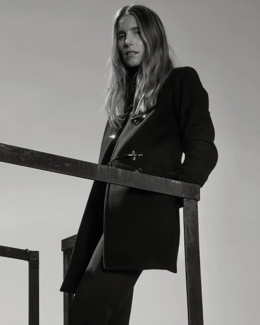 Monochrome portrait of an adult female in a dark coat standing on a black metal structure against a light backdrop in a fashion editorial style.
