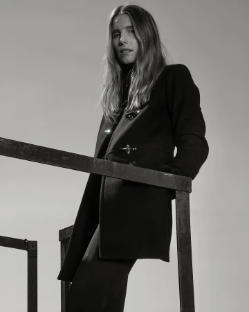 Monochrome portrait of an adult female in a dark coat standing on a black metal structure against a light backdrop in a fashion editorial style.