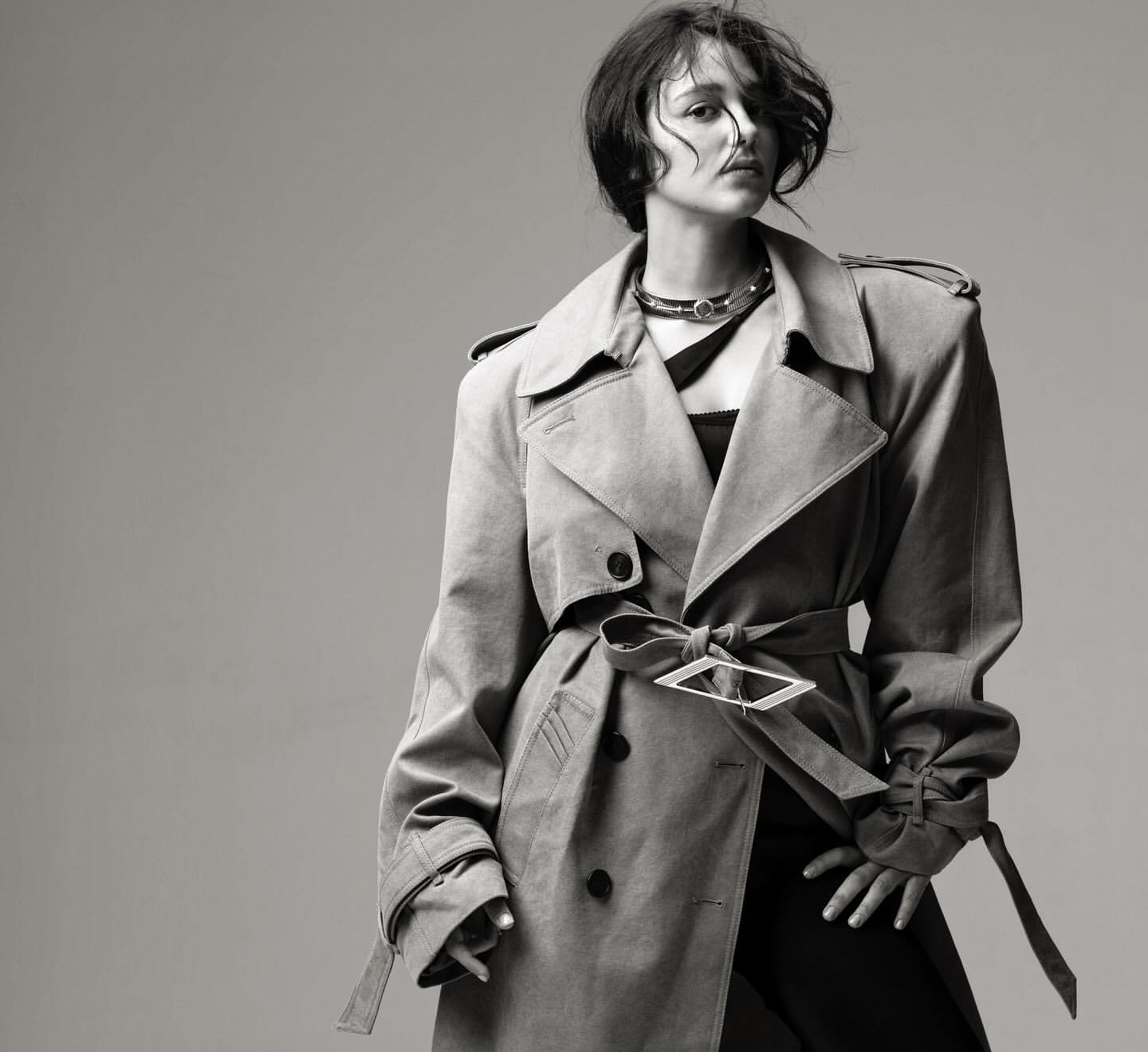 Black and white portrait of a stylish woman wearing a trench coat with a distinctive belt buckle and a choker against a gray backdrop.