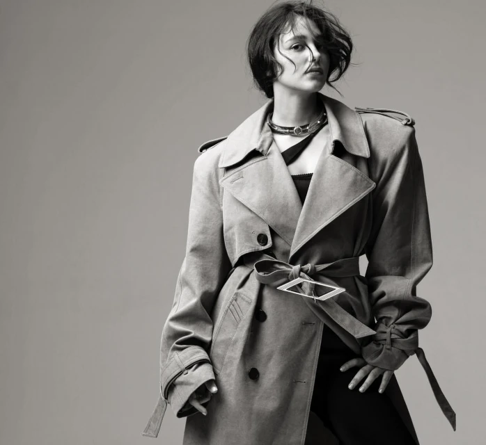 Black and white portrait of a stylish woman wearing a trench coat with a distinctive belt buckle and a choker against a gray backdrop.