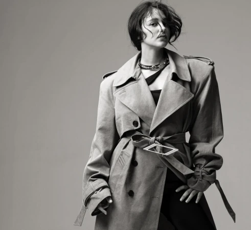 Black and white portrait of a stylish woman wearing a trench coat with a distinctive belt buckle and a choker against a gray backdrop.