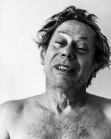 Black-and-white portrait of a shirtless adult male with tousled hair and eyes slightly closed, set against a plain background.