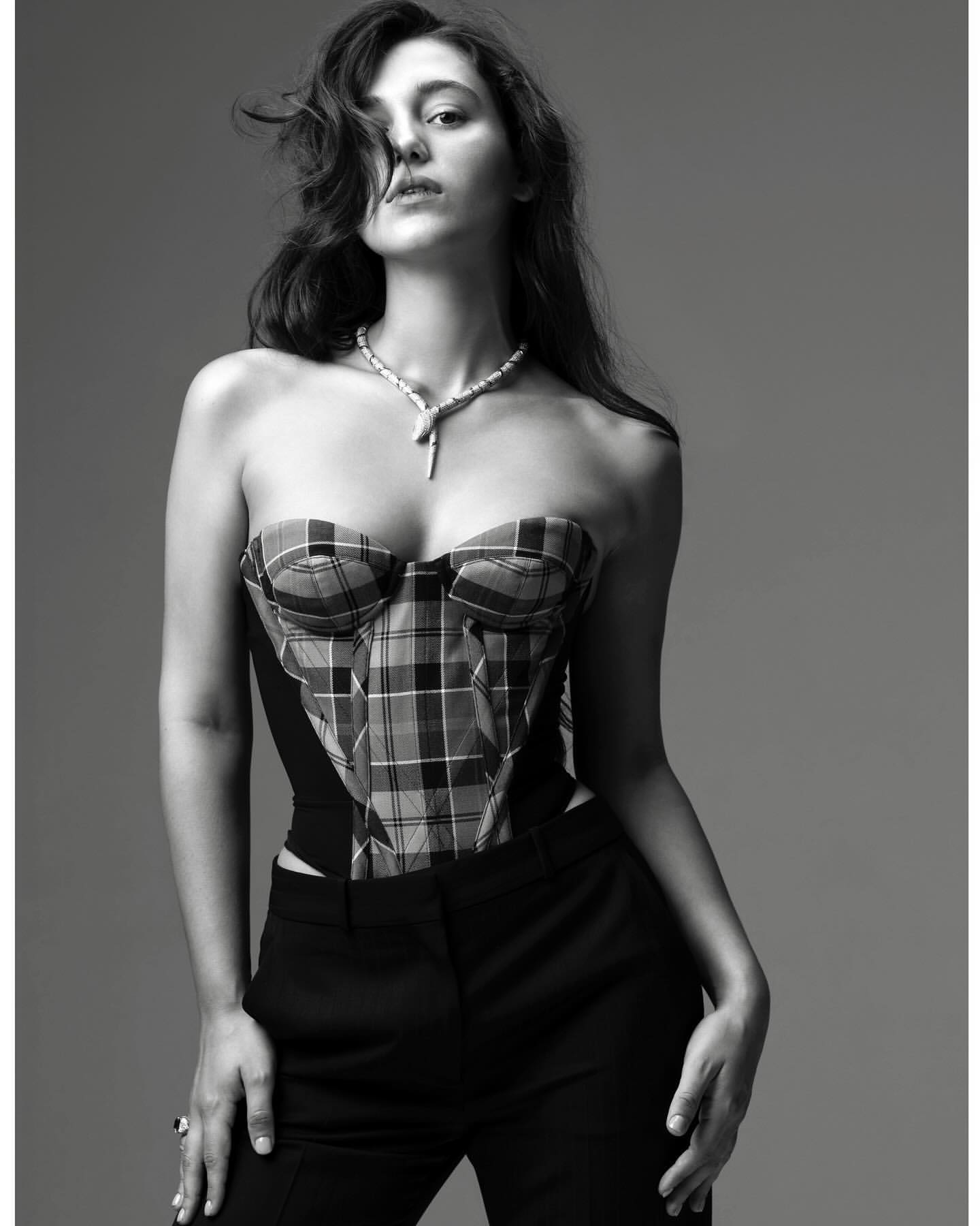 A woman poses in a black and white studio shot, wearing a plaid bustier and black pants, with a necklace, against a gray backdrop.