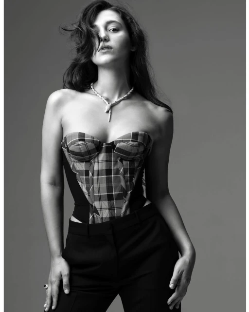 A woman poses in a black and white studio shot, wearing a plaid bustier and black pants, with a necklace, against a gray backdrop.
