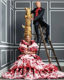 A woman in a dramatic red and pink gown seated below a man perched on a wooden ladder next to an towering sculpted blonde updo hairstyle.