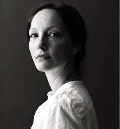 Black and white portrait of a woman in a white linen shirt looking back over her shoulder against a dark background