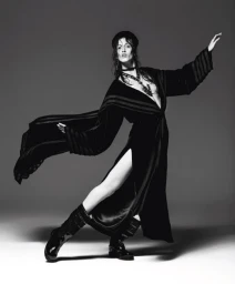Model in a black robe and boots striking a dynamic pose against a grey studio backdrop by Michael Avedon.