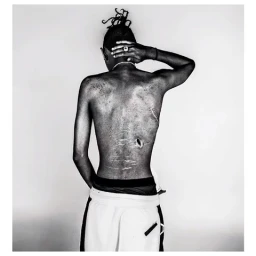 Black and white portrait of a shirtless man from behind with one hand on his neck, wearing light pants against a white background.
